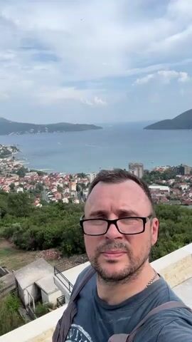 This is a selfie video taken by a man with dark hair, a beard, and glasses, wearing a dark blue t-shirt and grey overalls. He is standing on a terrace with a stone railing, looking directly at the camera with a neutral expression. Behind him, a scenic view unfolds of a coastal town nestled between green hills and a large bay.

The town features a cluster of red-roofed buildings, some taller, modern structures, and a coastline dotted with boats. The bay stretches out towards distant, hazy mountains under a partly cloudy sky. The sun appears to be shining, casting a bright light on the scene, suggesting it is daytime, likely late morning or early afternoon.

The man's forehead shows some redness, possibly from sunburn or an allergic reaction. A thin silver chain is visible around his neck. The overall mood is relaxed, with the man capturing a moment of enjoyment against a beautiful natural backdrop. The setting is clearly a coastal area, with the architecture and landscape indicative of a Mediterranean or Balkan region, specifically Sušćepan, Montenegro, as identified. There is some faint text on the man's t-shirt, but it is not legible.