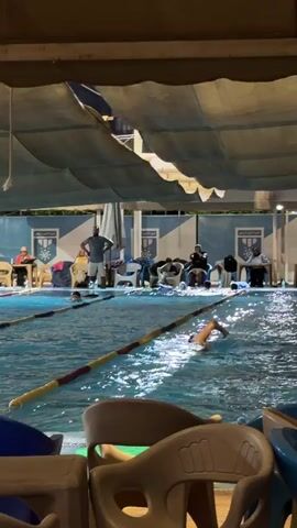 At a shaded swimming pool, several people are gathered around the poolside. Some are seated on chairs, observing those in the water. One individual in the water appears to be swimming, while another person standing by the pool's edge seems to be an instructor or coach, gesturing with their hands. The environment suggests an organized swimming activity or training session taking place during the evening, indicated by the artificial lighting.