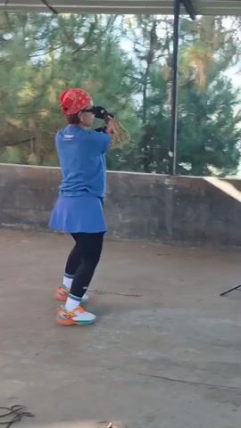 A woman with a red bandana and sunglasses is dancing on a concrete floor. She is wearing a blue t-shirt, a blue skirt, black leggings, and colorful sneakers. The woman is positioned in the center of the frame, facing left and moving her arms. Behind her, there is a black speaker on a stand, and a concrete wall. Trees are visible in the background. The scene appears to be outdoors under a covered structure.