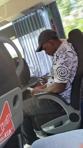 The video shows the interior of a bus with several passengers. In the foreground, a person wearing a black baseball cap and a patterned shirt is seated and looking at their phone. To the left of this person, a seatback tray table with a red triangle sticker with white text that says "KLIK" is visible. The bus has fabric seats and curtains covering the windows. The lighting suggests it is daytime.