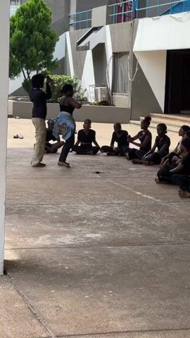 A group of people, mostly young women wearing black, are sitting on a concrete ground in an outdoor courtyard. Two individuals, one in a black top and khaki pants and the other in a black top and a denim skirt, are standing and appear to be engaged in a performance or rehearsal. The standing individuals are performing some kind of dance or movement with their arms. The background shows a building with a modern architectural design, trees, and outdoor seating areas. The lighting suggests it is daytime, possibly overcast.