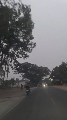 The video shows a POV shot from a moving vehicle on a road. The sky is overcast and the light suggests it is either early morning or late afternoon. On the left side of the road, there is a dirt path with some trees and a few buildings in the background. A person on a bicycle is visible on the dirt path, wearing a red shirt. Further down the road, a person is riding a motorcycle with a passenger. The road itself has some traffic, with headlights from oncoming vehicles visible. The overall scene depicts a rural or semi-urban setting on a cloudy day.