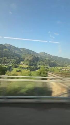 The video shows a view from a moving train window. The landscape outside is characterized by rolling green hills and mountains, dotted with scattered houses and trees. The sky is clear and blue with a few wispy clouds. In the foreground, a railing and blurred trees indicate the train's movement. There are no people visible in the frame. The scene is bathed in bright sunlight, suggesting it is daytime.