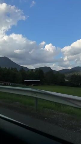 The video shows a landscape from inside a vehicle. A metal guardrail is visible in the foreground. Beyond the guardrail, there is a grassy area with trees and a small building. In the background, there are mountains under a blue sky with white clouds. The sun is shining.