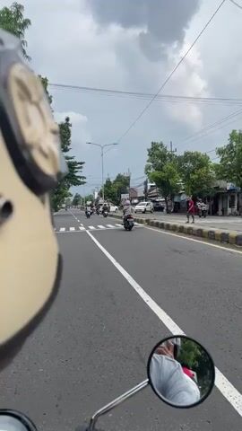 The video provides a first-person perspective from a motorcycle rider traveling along a road in Kertobanyon, Indonesia, under an overcast sky.

The top left of the frame shows a portion of a beige motorcycle helmet, while the bottom right features the motorcycle's rearview mirror, reflecting the rider's arm and torso, clad in a light-colored shirt.

The asphalt road is marked with clear white dashed and solid lines, as well as a prominent zebra crossing at the initial moments of the clip. Traffic primarily consists of other motorcycles and scooters, with multiple riders visible ahead and alongside the camera's perspective. A few cars are seen in the distance or parked along the road. At the beginning, a pedestrian in a red shirt is briefly visible walking on the right-hand sidewalk.

The sides of the road are lined with lush green trees, utility poles supporting numerous power lines, and a mix of single-story and multi-story buildings that appear to be residential and commercial. A street light pole and a traffic light are also visible further down the road.

The activity shows the rider moving forward with the flow of traffic. The sky is predominantly cloudy, indicating daytime conditions. A legible sign, briefly visible at 0:04, reads "AP AGRO."