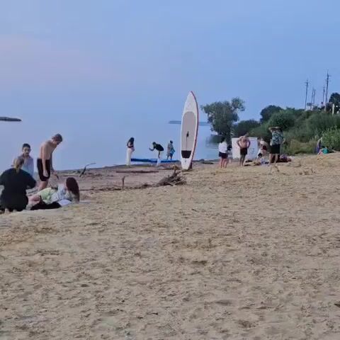A group of people are gathered on a sandy beach near a body of water. In the foreground, a few individuals are relaxing on the sand, with one person lying down and another sitting. Further back, a man is standing and appears to be taking a picture with his phone. A woman in a long white dress is also present. A red and white paddleboard is standing upright on the beach. Several other people are scattered on the beach, some standing and others sitting or lying down. In the background, there is water and a tree-covered shoreline. The sky is overcast and appears to be dusk.
