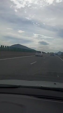 The video shows a view from inside a car driving on a highway. The highway has multiple lanes and is bordered by a green fence on the left and a grassy shoulder with some trees and hills on the right. The sky is cloudy with patches of blue peeking through. In the distance, there are mountains. Several cars are visible on the highway, including a white SUV with a spare tire on the back, a black SUV, and a few other sedans. The time of day appears to be daytime, given the brightness of the sky, and the weather is partly cloudy. The license plate of the white SUV is visible and appears to be "E989FC 13". The overall scene conveys a sense of travel and journeying through a somewhat rural or mountainous landscape. The location is identified as Konstantinovskaya, Russia.