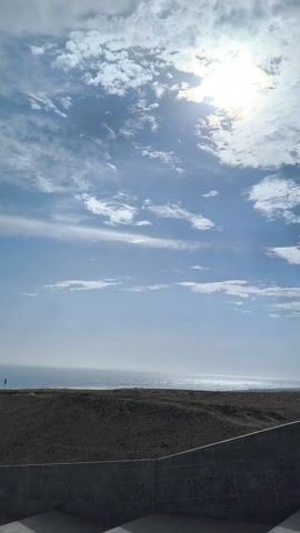 The video presents a wide shot of a beach scene beneath a sky filled with white clouds and a bright sun. The scene is captured from an elevated position, potentially a staircase or a viewing platform, as indicated by the concrete barrier in the foreground.  Below, a sandy dune slopes down towards a beach bordered by the ocean. The ocean stretches across the horizon, with waves breaking near the shore.  Adjacent to the dune, a concrete structure or series of structures and pathways runs parallel to the beach.  The overall scene conveys a sense of calm and natural beauty.