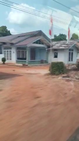 The video shows a view from inside a moving vehicle looking out at a rural landscape. The scene features several small, single-story houses with weathered roofs. The houses are white or light-colored and appear to be simple structures. In front of some of the houses are manicured bushes and trees, while other areas show bare, reddish-brown earth. Power lines are visible crisscrossing the sky above. In one section, an Indonesian flag is flying on a flagpole. The perspective suggests the viewer is passing by these houses, with some motion blur indicating the vehicle's movement. The overall environment is a tropical or subtropical setting, characterized by the vegetation and the style of the houses.