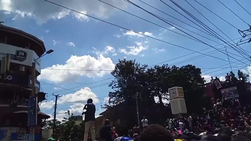 Here is a description of the video:

The video shows a large crowd gathered in what appears to be a city street. The crowd is dense and mostly comprised of people with dark hair. Many are wearing casual clothing and hats. A man in a suit stands on top of a vehicle, possibly a car or truck, addressing the crowd. He is speaking into a microphone. Behind the crowd and the speaker, there is a truck or platform elevated above the crowd, with people standing on it. There are also large speakers on the platform. The background features buildings with commercial signs, power lines crisscrossing the sky, and some trees. The sky is bright blue with scattered clouds. The overall impression is that this is a political rally or public gathering.