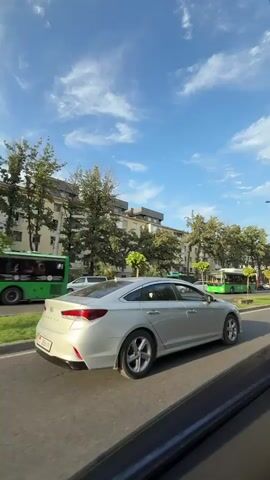 This video shows a view from inside a moving vehicle, looking out onto a city street. The focus is on a silver sedan driving in the right lane, with its rear end prominently displayed. The car has a license plate that is partially visible. To the left of the sedan is a green bus. Further to the left, on the sidewalk, are several parked cars and buildings, some of which appear to be apartment complexes with trees in front of them. A large red truck with the words "MÖBEL SCHOTT" on its side is also visible in the distance. The sky is blue with white clouds. The road is asphalt and appears to be in good condition. The scene is lit by daylight, suggesting it is daytime.