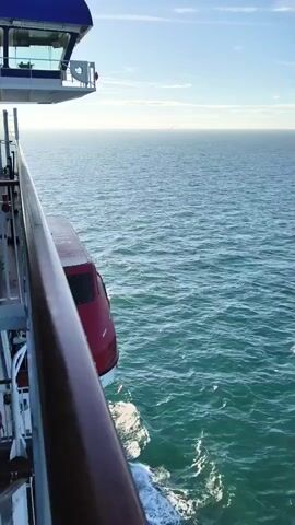 The video shows a view from the side of a large vessel sailing on a calm, blue-green ocean under a clear sky. A red lifeboat is attached to the side of the vessel, hanging just above the water. The vessel appears to be moving, as evidenced by the wake trailing behind it and the slight tilt of the lifeboat. The sun is low in the sky, casting a warm light on the scene.