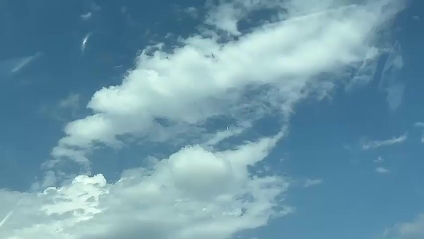 The video shows a view of a bright blue sky filled with scattered white clouds. Some of the clouds are fluffy and voluminous, while others are wispy and streaky. There are also some faint linear contrails visible in the sky, suggesting the passage of aircraft. The view appears to be from a vehicle, as there are reflections on the surface of the glass in the foreground, including what looks like the edge of a windshield wiper and the curve of a car's interior. The sunlight is strong, illuminating the clouds and casting a bright light on the scene.
