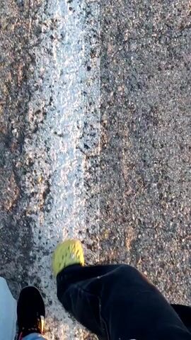 The video shows a point-of-view perspective walking on an asphalt road with a white line down the center. The road is cracked and weathered. Tall grass is on the left side of the road. In the distance, a car is parked on the side of the road. The sky is blue with some clouds, and the sun is setting, casting long shadows and a warm light. The camera is pointed downwards at the feet of two people walking. One person is wearing black shoes with pink and white accents, and light blue pants. The other person is wearing yellow Crocs with embellishments and dark pants. The person in the yellow Crocs is slightly ahead and to the right of the person in the black shoes. The camera pans and tilts slightly, capturing the movement of the walkers and the environment around them.