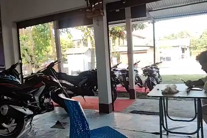 The video shows the interior of a motorcycle showroom. In the foreground, a person's face is partially visible as they look down. In the background, several motorcycles are displayed. To the right, a man sits at a small table, looking at his phone. The environment appears to be a covered outdoor area, possibly a dealership's front display. The lighting is natural, suggesting daylight.
