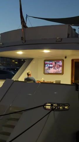 A man is sitting facing away from the camera on a yacht, looking at a television screen. The television screen shows two people, one male and one female, in a room with shelves of products behind them. The man on the yacht is wearing a blue shirt and has gray hair. The television screen is mounted on a white wall. The yacht appears to be docked at night, with lights visible in the background.
