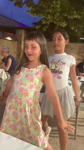 Two young girls are dancing outdoors at dusk. The girl in the foreground wears a floral print dress and has her arms outstretched. The girl behind her wears a white t-shirt and a grey skirt, also with her arms raised. They are at a restaurant, with tables and chairs visible in the background. The lighting suggests it is evening.