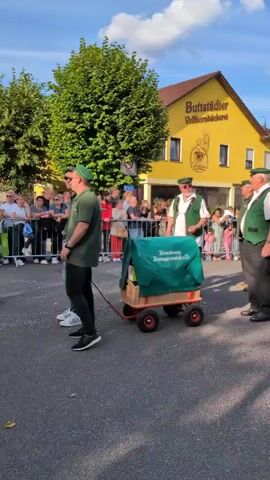 The video shows a parade or festive procession in a town. In the foreground, three men in matching green vests and hats, along with a man in a green shirt, are walking. They are pulling a small red wagon covered with a green tarp that has text on it. Behind them, a crowd of spectators stands behind a black metal fence. In the background, a yellow building with a sign that reads "Buttstädter Vollkornbäckerei" is visible, along with other buildings and trees. The sky is blue with some white clouds. The men in vests are waving to the crowd. The overall atmosphere appears to be celebratory.