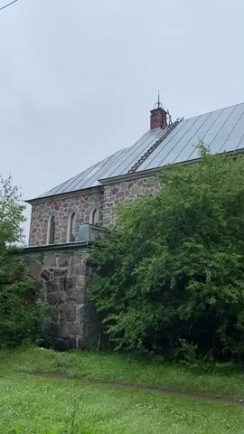 The video shows a close-up, exterior view of a stone building, possibly a church, with a metal roof. The building is covered in a rough stone masonry, with arched windows visible on the upper portion of the facade. A dense green tree or bush obscures a significant portion of the building from view in the foreground. The sky is overcast, suggesting a cloudy day. The camera pans slowly from left to right, revealing more of the building's structure and the surrounding vegetation. The scene appears to be in a rural or semi-rural setting, with a grassy area in the foreground. There are no people visible in the video.
