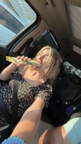 A blonde woman wearing a floral dress is sitting in the passenger seat of a car. She is holding a wrapped food item and taking a bite out of it. The car appears to be in motion as the scenery outside the window is blurred. The interior of the car is visible, including the seatbelt, dashboard, and a portion of the driver's arm. Sunlight is streaming into the car.