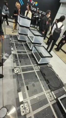 The video captures an indoor scene at what appears to be an exhibition or trade show, given the presence of attendees and displays. The camera is positioned at a high angle, offering a detailed view of a gray and black grid-like structure on the floor. The grid is composed of square tiles with various details and connection points visible. Several transparent plastic storage containers are placed on the grid in a staggered formation. An arm and hand of a person are visible, pointing at a specific section of the grid. Background activity includes multiple people walking around, some wearing masks, and signage with the words "Tokyo Future Tour 2035" is visible. The general atmosphere suggests an event centered on technology or future innovations.