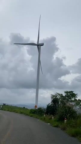 This video shows a series of scenic views of wind turbines set against a cloudy sky. The first frame features a single, tall wind turbine positioned next to a road with a warning sign at its base. The second frame captures two wind turbines in the distance, partially obscured by tall grasses in the foreground. The third frame offers a wider view of the landscape, with two wind turbines on a grassy hill and lush greenery surrounding them. The fourth frame presents a panoramic view of rolling hills and mountains under a heavy cloud cover, with a large tree dominating the right side of the image. The final frame returns to a view of a wind turbine on a grassy hill, with distant mountains and clouds forming the backdrop. There are no people visible in any of the frames.