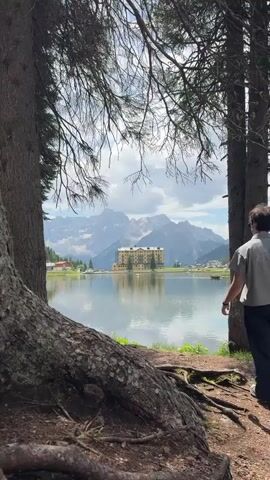 This media file captures a serene landscape scene featuring a person looking out at a picturesque view. The primary subject is a person standing with their back to the camera, positioned on the right side of the frame. They are wearing a light-colored, short-sleeved shirt and dark trousers. The person is looking towards a large, tranquil lake that dominates the mid-ground of the image.

The setting is a natural environment, with a forest framing the view. Two large trees are prominently featured in the foreground on the left and right, with their branches extending overhead, creating a natural archway. The ground in the foreground is covered in dirt and exposed tree roots, suggesting a wooded area.

Across the lake, a large, light-colored building with a distinctive architectural style stands out. It appears to be a hotel or a grand establishment, reflected in the still waters of the lake. Behind the building and the lake, majestic, rugged mountains rise sharply against a partly cloudy sky. The mountains are snow-capped in some areas, indicating a high altitude and possibly a cooler climate. Small buildings are visible on the shore of the lake to the left of the main building.

The activity taking place is one of contemplation and observation. The person is standing still, seemingly taking in the breathtaking scenery. Their posture, with one hand raised slightly to shield their eyes from the sun, suggests they are admiring the view and possibly the mountains in the distance. The overall mood conveyed is one of peace, awe, and appreciation for nature's beauty.

The time of day appears to be daytime, given the bright lighting and the clear visibility of the landscape. The weather seems pleasant, with a mix of sun and clouds in the sky, contributing to the scenic beauty. The location is strongly suggested to be Auronzo di Cadore, Italy, known for its stunning alpine lakes and mountains, consistent with the visual elements in the media. There is no visible text in the media file.