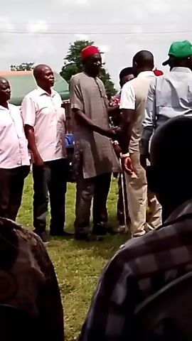 A man in a grey tunic and pants and a red cap is shaking hands with another man. The second man is wearing a white shirt and beige pants. Several other men are standing around them, some looking at the interaction. The scene appears to be outdoors, possibly a gathering or event, with grass and trees visible in the background, along with some tents. The overall lighting suggests daytime.