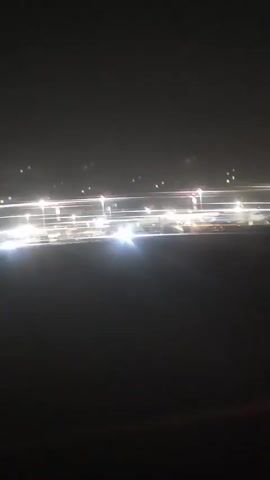 The video shows a blurry, dark night scene from the perspective of a moving vehicle, likely an airplane on a runway. Bright lights from airport infrastructure, including what appear to be runway lights and building lights, create streaks across the frame. Several airplanes are visible, parked on the tarmac. Some of these airplanes have stairs attached, suggesting passengers may be boarding or disembarking. The overall impression is of a busy airport at night.