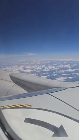 An aerial view from an airplane window shows a vast expanse of clouds under a clear blue sky. The wing of the airplane is visible in the foreground, dominating the left side of the frame. Below the clouds, a landscape of hills and some scattered buildings can be discerned, suggesting a rural or sparsely populated area. The clouds are fluffy and white, with some appearing denser and casting shadows. The sunlight illuminates the scene, creating a bright and serene atmosphere.