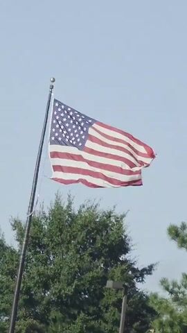 An American flag on a flagpole is shown against a clear blue sky. The flag is partially unfurled and appears to be waving in the wind. In the background, the tops of green trees are visible, and a light-colored pole with a dark fixture is partially visible in the lower right corner.