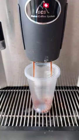 A close-up shot shows an SCS (Swiss Coffee System) machine dispensing coffee into a clear, plastic cup. The machine is black with a stainless steel front and a logo featuring the Swiss flag and the text "Swiss Coffee System." The coffee being dispensed is a dark amber color. The cup sits on a metal grate.