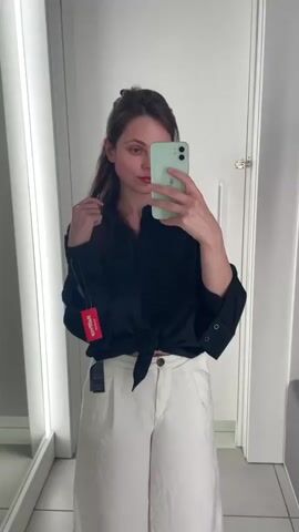 A woman stands in a fitting room, taking a selfie with her phone. She is wearing a black tied-front shirt and white wide-leg pants. The fitting room has mirrors on the left wall and a door on the right. There are clothing tags visible on her shirt.