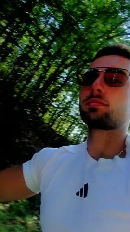 This media file captures a first-person perspective of riding an all-terrain vehicle (ATV) on a paved road surrounded by dense, green foliage. The rider, wearing a white t-shirt, is visible in the ATV's rearview mirror, as is the passenger in the first frame. The ATV's handlebars, instrument panel, and handguards are prominent in the foreground. The road appears to be a rural path, winding through a wooded area with tall trees and lush undergrowth. Sunlight filters through the leaves, creating dappled patterns on the road and indicating it is daytime, likely late morning or afternoon. The atmosphere appears peaceful and leisurely, suggesting a recreational ride. No text is visible within the video itself. The context provided indicates the location is Orlovtsi, Bulgaria.
