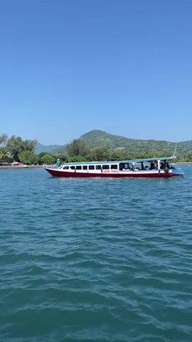 The video shows a long, red and white boat moving on the surface of calm, turquoise water under a clear, blue sky. The boat, labeled "AJAR PAGI", has several people on board. In the background, there is a green shoreline with trees and a few buildings, as well as a large hill or small mountain in the distance. The camera is positioned at water level, providing a steady, wide shot of the boat as it travels across the water. The overall scene is bright and sunny, suggesting a clear day at sea or on a large lake.