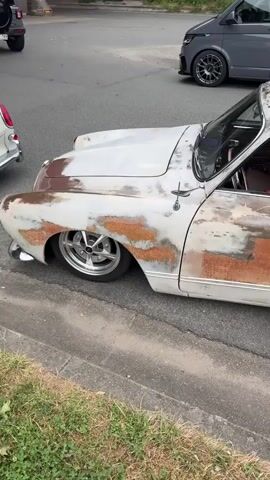 The video shows a vintage car with a weathered, rusty paint job parked on the side of a paved road next to a grassy area. The car is white with large patches of rust, and it has chrome wheels. The car is low to the ground, suggesting it has been modified. In the background, a gray van with a logo and a portion of another white vehicle are visible. The scene appears to be outdoors during the daytime.