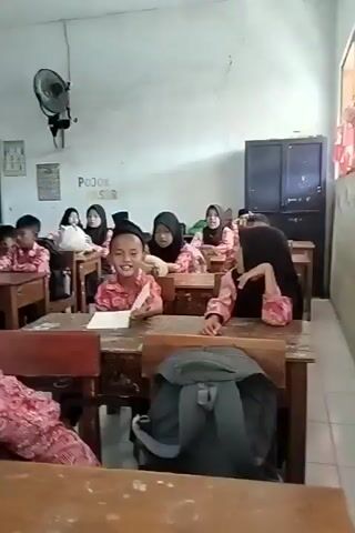 The video shows a classroom filled with young students, most of whom are wearing matching pink and brown patterned shirts and some wearing black peci caps. They are seated at wooden desks, facing towards the front of the classroom. There's a green board visible on the wall, and a black ceiling fan mounted above. Some students are looking at the camera, while others appear engaged with something in front of them, possibly an activity or lesson. One student in the foreground is leaning on their desk. The overall environment suggests a school setting, possibly during a class or an event.