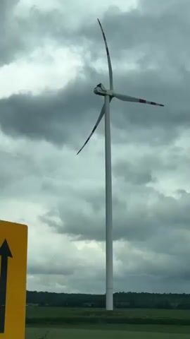 The video shows a tall, white wind turbine with red-tipped blades against a background of dark, cloudy skies. The turbine stands in a green field with a treeline in the distance. The camera appears to be moving, likely from a car, as a tree briefly obstructs the view. The turbine blades are turning slowly.