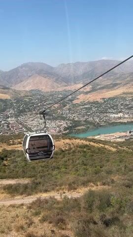 The video shows a view from a cable car cabin moving over a hilly, semi-arid landscape. Below, a city is visible, with buildings and streets spread out. A bright blue body of water, likely a lake or reservoir, is also present in the middle distance, with some industrial or dock-like structures along its edge. Further in the background, a range of dry, rugged mountains under a clear blue sky. The cable car itself is white and modern-looking, with darkened windows. Other cable car cabins can be seen further down the line, also ascending the slope. The camera is positioned inside the front-facing cabin, looking out and slightly down at the panoramic view. The overall impression is one of scenic travel over a populated area nestled in a mountainous region.