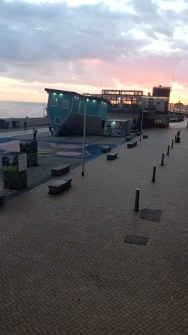 The scene is a seaside promenade at dusk or dawn. The sky is partly cloudy with the sun creating a colorful gradient on the horizon. To the left, a sandy beach meets the ocean. Several people are walking or standing on the beach. The promenade itself is paved with light brown bricks and features benches and decorative pillars. Some pillars are clad in rock and display photographs. A blue building, designed to appear upside down, stands to the right of the frame. Further to the right, another building can be seen in the background. The camera pans slightly to the left, showing more of the beach and the arrangement of the benches and pillars on the promenade.
