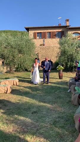 The video captures a wedding procession in Montepulciano, Italy. A bride in a flowing white gown, with a deep V-neck and a thigh-high slit, walks arm-in-arm with an older man in a dark suit, white shirt, and blue tie. The bride carries a bouquet of white flowers. They are walking down a grassy path lined with large, burlap-covered cushions or seating, suggesting an outdoor ceremony. Olive trees with full green foliage frame the scene, and in the background, a rustic stone building with red-tiled roofs stands under a clear blue sky. The sunlight suggests it is late afternoon or early evening, casting long shadows on the grass. The overall mood appears joyful and celebratory, as indicated by the smiles of the bride and her escort, and the presence of guests in the background. Other guests are visible, some seated on the cushions, and others standing near the building. One woman in a blue jacket is clapping enthusiastically on the left side of the frame. A wooden barrel with a floral arrangement stands near the building, adding to the festive atmosphere. The architecture of the building and the olive trees strongly suggest a Tuscan setting. There is no visible text in the media.