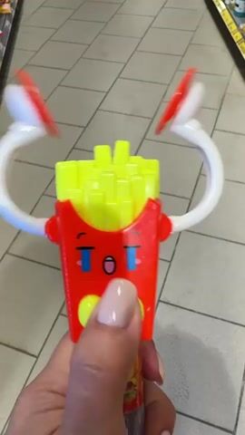 A close-up shot shows a hand holding a toy that resembles a carton of french fries. The toy has a red body with a yellow top designed to look like french fries. It features a cartoon face with blue tear streaks, an open mouth, and closed eyes with eyelashes. Two yellow circles represent cheeks, and a yellow bow tie is located below the mouth. The toy has two white curved arms with red disc-shaped attachments on the ends. These arms are in a position that suggests they are raised. The background appears to be a tiled floor in what looks like a store aisle, with shelving visible in the upper portion of the frame. The hand holding the toy has painted fingernails in a neutral shade.