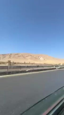 The video shows a landscape seen from a moving vehicle. In the foreground is an asphalt road with concrete barriers. In the background is a desert landscape with a low mountain range under a clear, blue sky. The vegetation is sparse, mainly consisting of small trees and shrubs. As the vehicle moves, it passes by a decorative concrete wall.