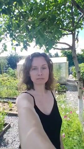 A woman with curly brown hair, wearing a black camisole, stands outdoors. She is holding a camera and taking a selfie. In the background, there is a garden with green plants, flowers, a greenhouse, and some buildings. The sky is blue with scattered clouds. The camera pans slightly, showing different angles of the garden and the woman.