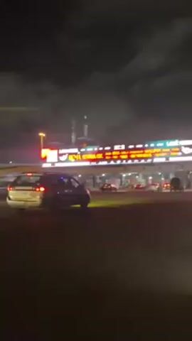 The video shows a toll plaza at night, with cars passing through the booths. There is a large electronic billboard above the toll booths displaying advertisements. The sky is cloudy and the area is illuminated by artificial lighting.