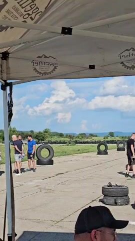 Under a white tent, a group of people and cars are on a paved area with a grassy field and hills in the background under a blue sky with white clouds. On the left, a dark green car with its hood open is attended by a man in a white t-shirt and grey shorts, who is leaning into the engine bay. Another man in a black t-shirt and white shorts is standing near the trunk of the car. To the right of the green car, a group of three men are standing and talking. One man is wearing a light blue t-shirt and dark shorts, another is in a grey t-shirt and black shorts, and the third is in a black t-shirt and black shorts. Several black tires are stacked or laid on the ground near the group and further down the paved area, which appears to be a track or staging area. In the distance, a man in a black t-shirt and black shorts is walking towards a silver car with its hood also open, near more stacked tires. The tent overhead has upside-down text and logos visible, indicating it's a branded canopy.