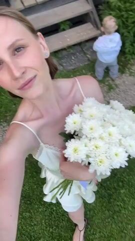 A young woman in a white satin slip dress smiles as she holds a bouquet of white chrysanthemums. She's standing on a green lawn, and there's a stone path visible in the background. Someone walks behind her briefly. The woman appears to be taking a selfie, as the camera is pointed at her. She’s wearing a small earring.