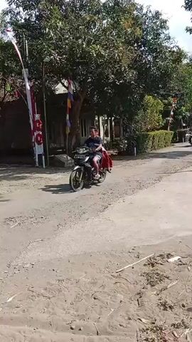 A person rides a motorcycle on a dirt road. The motorcycle has a man riding it and another person sitting behind him. There are trees and buildings in the background.