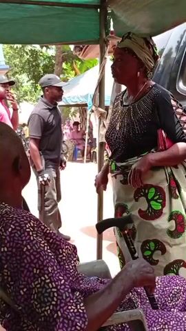 The video shows a gathering of people, likely outdoors under a green tarp, with a man in a wheelchair facing away from the camera. A woman wearing a colorful patterned skirt and headwrap is speaking to someone off-camera, holding a walking stick. In the background, other people are moving around, some wearing casual clothes, while others are dressed in more formal attire, including traditional African prints. There are speakers visible, suggesting there may be music or a performance. The setting appears to be in a residential area with trees and a building in the background.