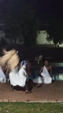 At night, against a backdrop of greenery and a pool, a dark-skinned person wearing traditional garb, including a straw-like head covering, a white cloth wrap, and ankle bells, is dancing with a stick. Two people seated nearby are also wrapped in white cloth, and others can be seen in the background.